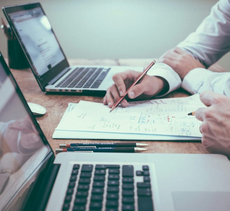 Zwei Personen arbeiten an Laptops und notieren Informationen auf Papier.